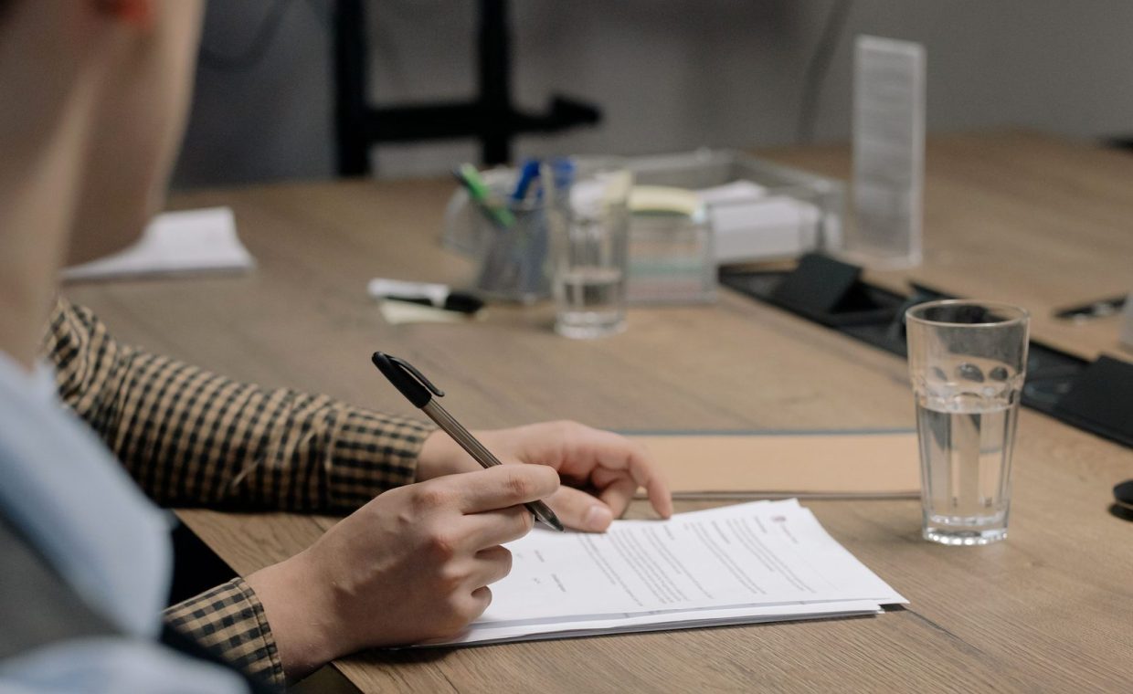 Person in business attire signing a document at a wooden table in an office setting.