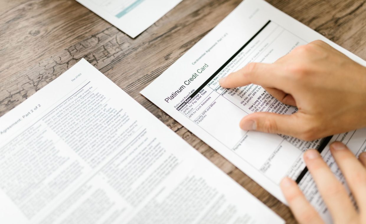 A hand examining a credit card agreement on a wooden desk, highlighting financial review.