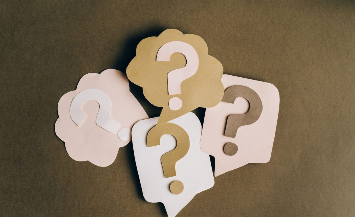Four paper cutouts of question marks in beige and white on a brown surface, symbolizing inquiry and curiosity.