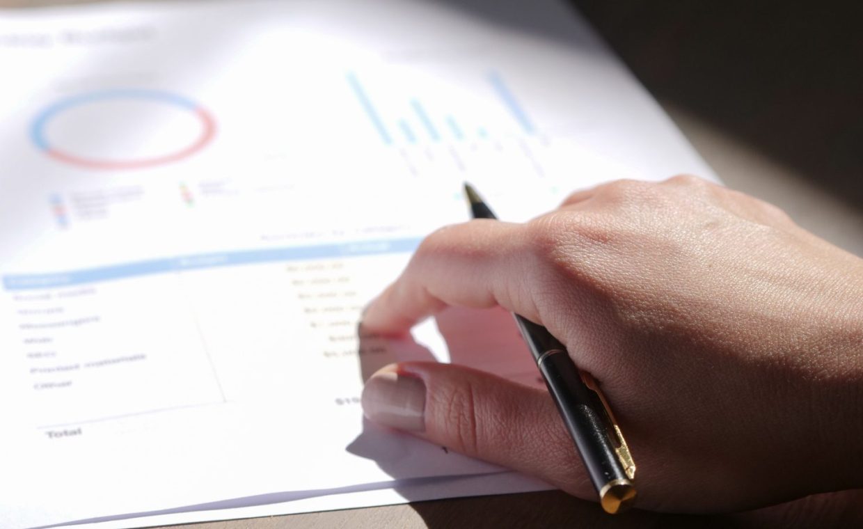 A close-up view of a hand holding a pen and examining financial charts on paper.