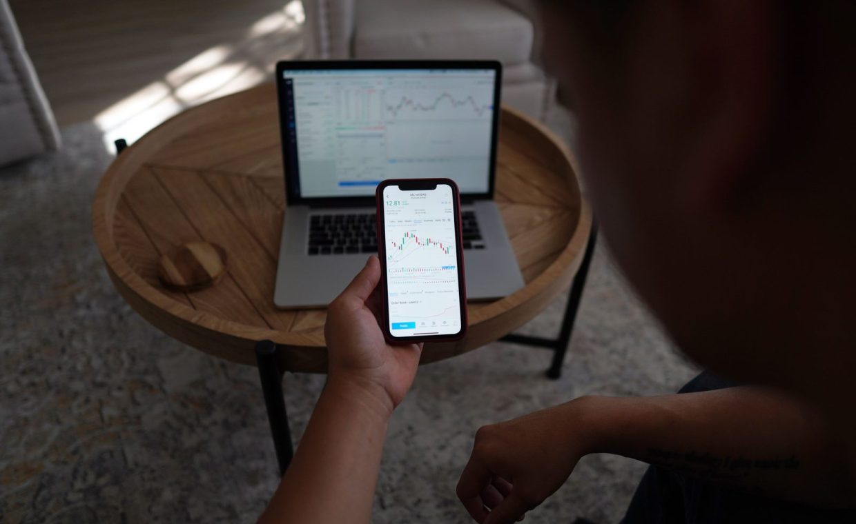 A person examines stock market graphs on a smartphone and laptop at home, showcasing remote financial analysis.