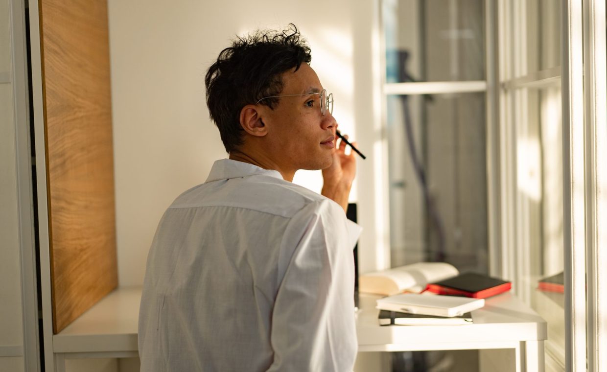 Man in white shirt gazing out window pensively, surrounded by books in modern office.