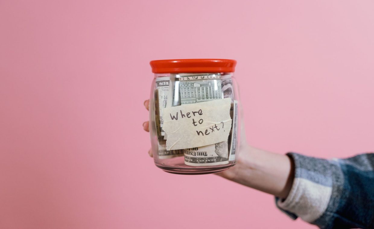A hand holding a glass jar filled with dollar bills labeled 'Where to next?' against a pink background, symbolizing savi