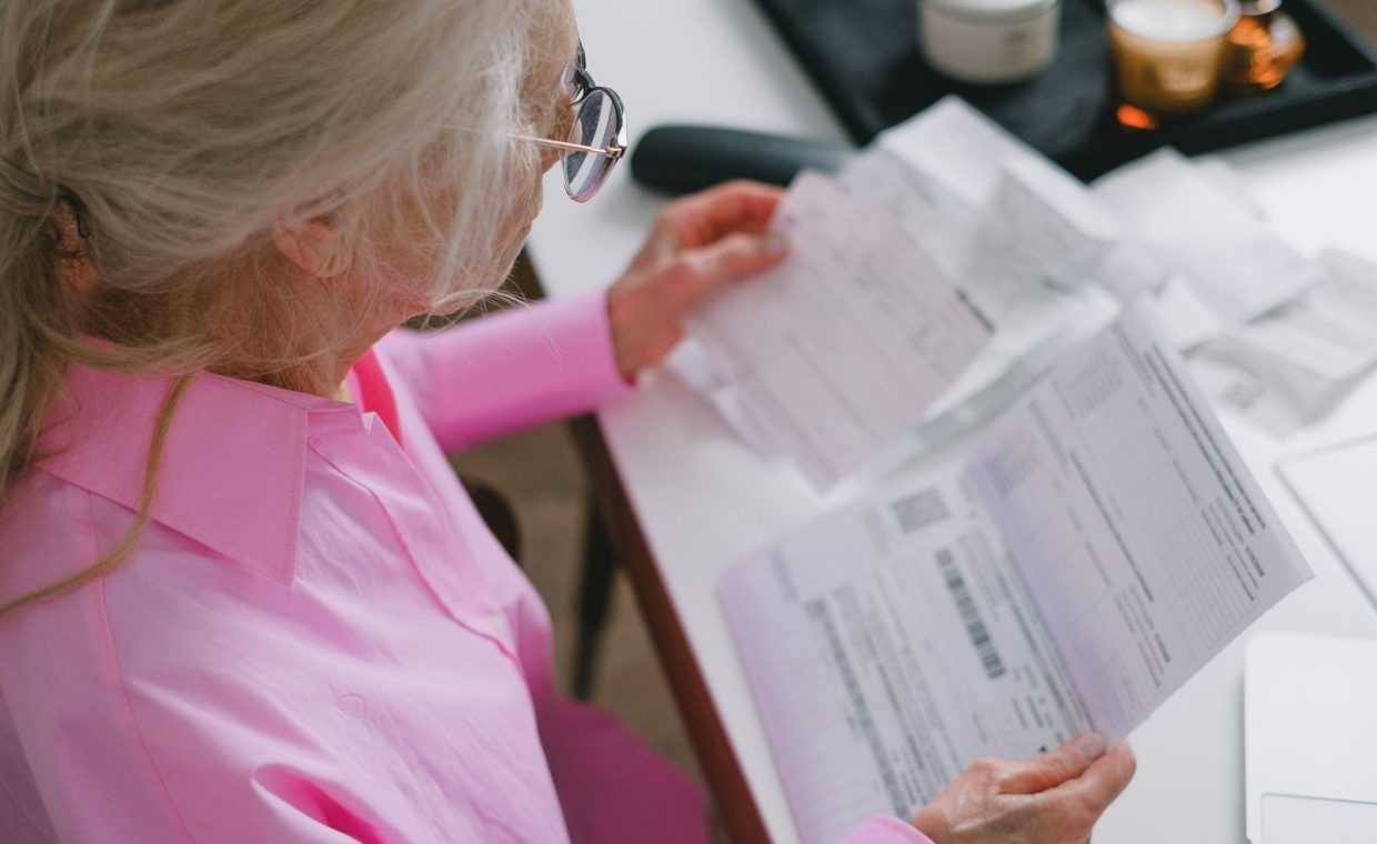 An elderly woman in glasses holds and reads important papers at a table indoors.