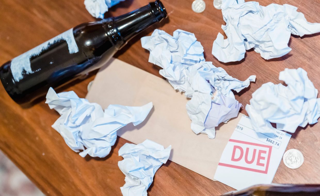 A chaotic scene with overdue bill, beer bottles, and crumpled papers on a wooden table.